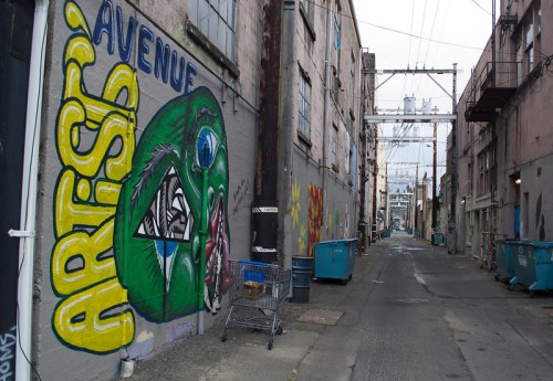 A narrow alley with colorful graffiti on the wall, featuring a green face and the words "Artists' Avenue." Trash bins are visible.