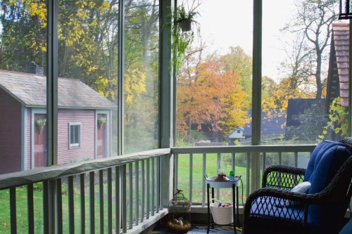 A cozy screened porch with a blue chair, overlooking a yard with autumn trees and a red shed in the background.