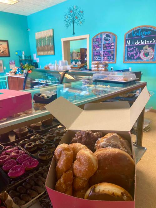 A box of assorted donuts in a colorful bakery with a display case and chalkboard menus in the background.