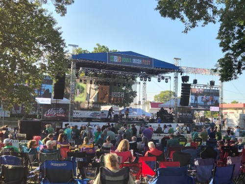 A crowd enjoys the Iowa Irish Fest, with a stage set up for performances under a clear blue sky.
