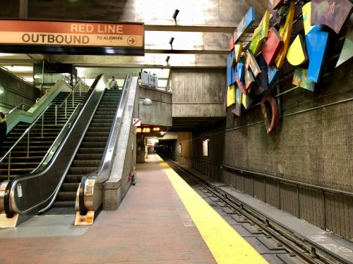 A subway station platform with escalators, colorful wall art, and empty tracks under a sign for the Red Line to Alewife.