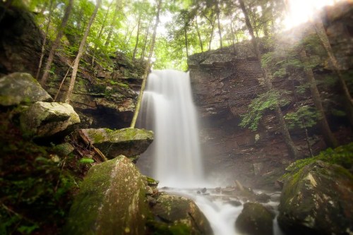 A serene waterfall cascades down rocky cliffs, surrounded by lush green trees and mist in a tranquil forest setting.