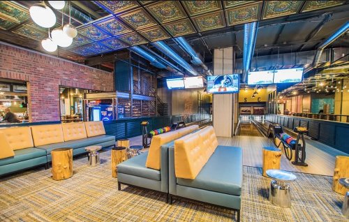 Modern lounge area with stylish seating, wooden accents, and a bowling alley in the background. Bright and inviting atmosphere.