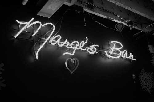 Neon sign reading "Marge's Bar" with a heart design, set against a dark background.