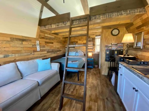 Cozy interior of a small cabin with a loft, featuring a sofa, bed, wooden walls, and a ladder.