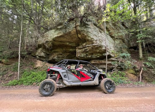 A person sits in a red and black off-road vehicle parked on a dirt road near a large rock formation in a forest.