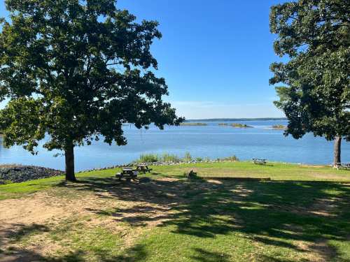 A serene lakeside view with trees, picnic tables, and calm water under a clear blue sky.