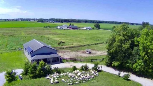 Aerial view of a farm with a barn, green fields, and a few horses in the distance under a clear blue sky.