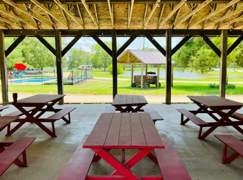 A covered picnic area with red tables, overlooking a pool and grassy area by a lake.