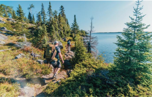 Two hikers trek along a rocky path by a lake, surrounded by lush green trees and a clear blue sky.
