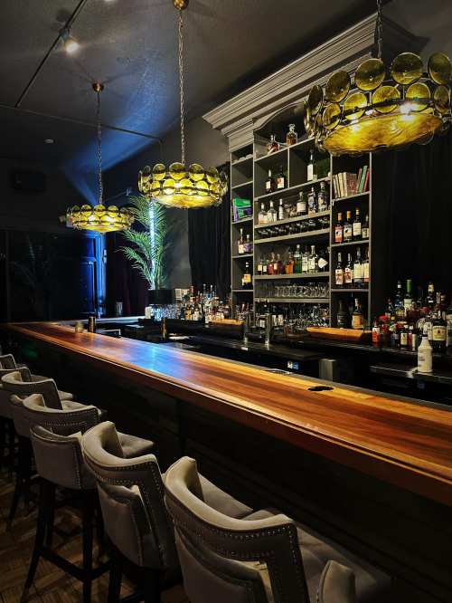 A stylish bar with a wooden counter, elegant seating, and unique pendant lights, surrounded by shelves of liquor.