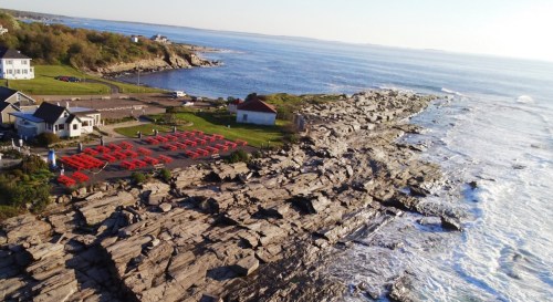 A coastal view featuring rocky shoreline, ocean waves, and a parking area with red chairs near a white building.
