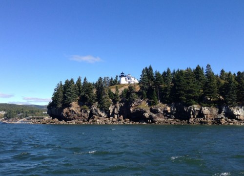 A lighthouse perched on a rocky cliff surrounded by trees, with calm waters in the foreground and a clear blue sky.