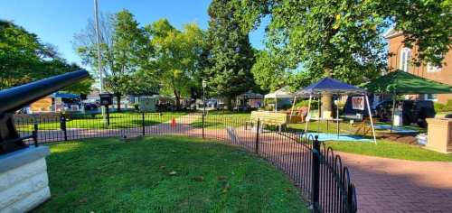A park scene with tents, trees, and a cannon, set up for an event with vehicles in the background.