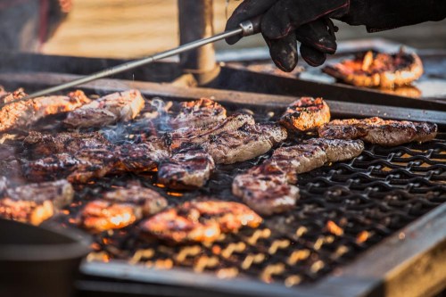 A close-up of grilled meat sizzling on a barbecue, with smoke rising and a hand using a spatula to flip the steaks.