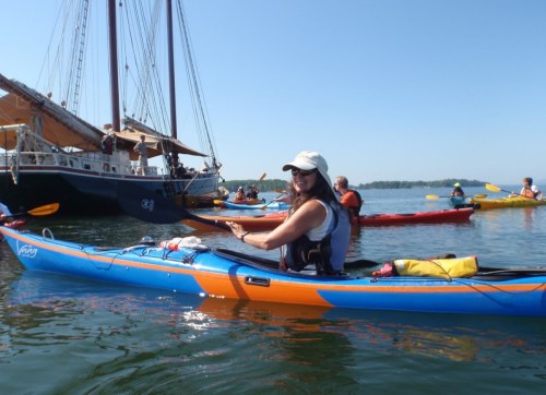 A woman in a kayak smiles while surrounded by other kayakers and a large sailing ship on calm waters.