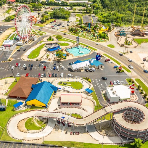 Aerial view of an amusement park featuring a Ferris wheel, roller coaster, and colorful buildings with parking lots.