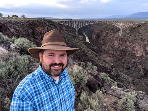 A man in a plaid shirt and hat stands on a rocky ledge with a canyon and bridge in the background.