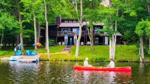 Two people paddle a red kayak on a calm lake, with a wooden house and trees in the background.