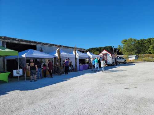 A sunny outdoor market scene with tents, vendors, and visitors in a gravel area near a rustic building.