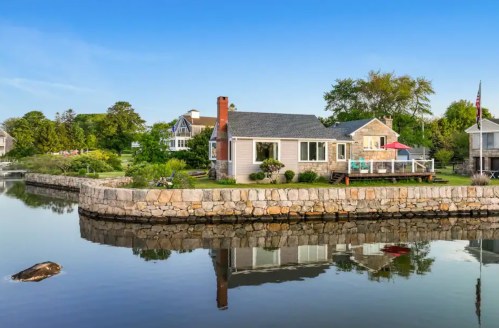 A charming waterfront cottage with a stone wall, surrounded by greenery and reflecting in calm water.