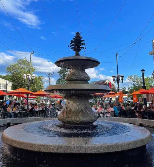 A decorative fountain in a lively outdoor dining area with umbrellas and people enjoying their meals under a blue sky.