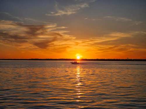 A serene sunset over calm waters, with a small boat silhouetted against the vibrant orange and yellow sky.