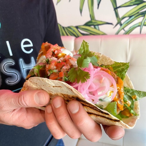 A person holds a taco filled with lettuce, salsa, and creamy sauce, set against a colorful background.