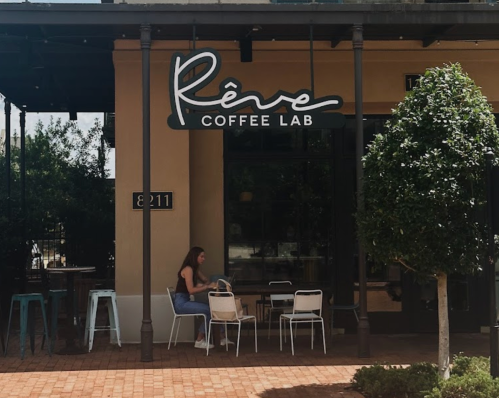 A woman sits at a small table outside Rêve Coffee Lab, with greenery and modern seating in a sunny urban setting.