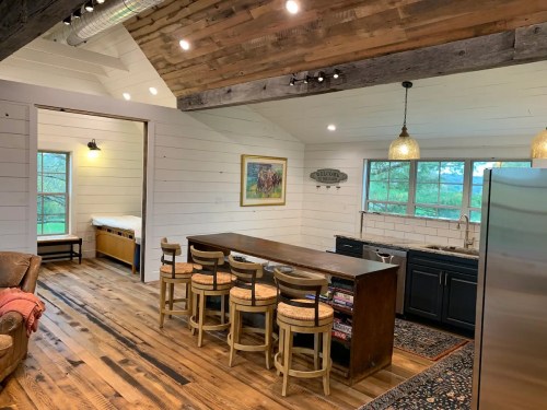 Cozy kitchen with wooden bar, stools, and large windows, featuring rustic decor and warm lighting.
