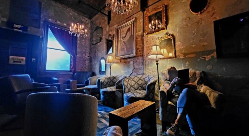 Cozy, dimly lit lounge with vintage decor, chandeliers, and a person sitting on a sofa, absorbed in their phone.