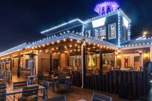 A brightly lit restaurant exterior at night, featuring string lights and outdoor seating on a wooden deck.