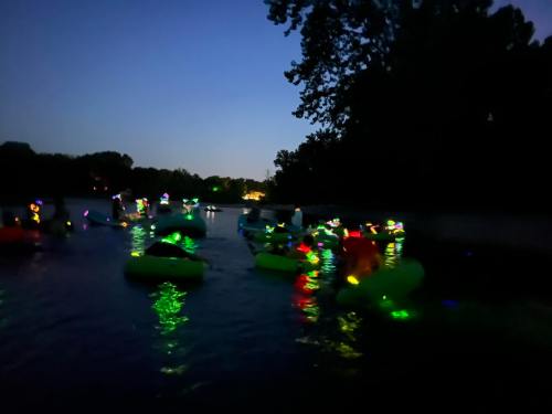A group of people floating on colorful, illuminated rafts in a dark river at dusk. Trees line the shore.