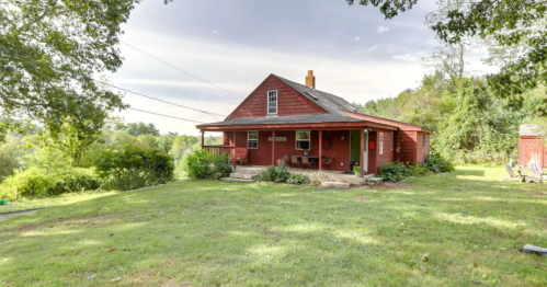A red, single-story house with a porch, surrounded by green grass and trees under a cloudy sky.