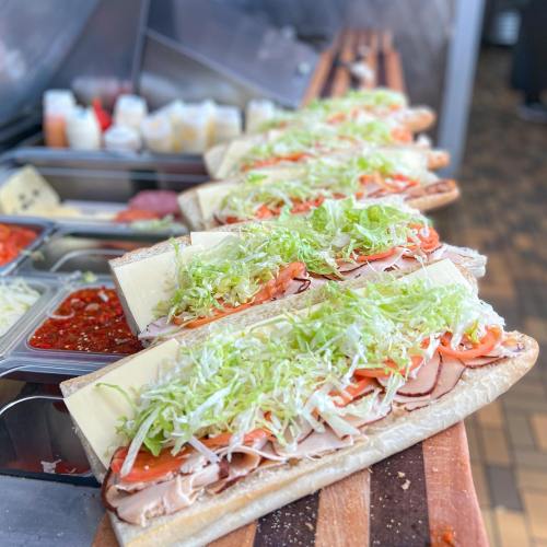 A row of large sandwiches filled with lettuce, tomatoes, and deli meats on a wooden counter.