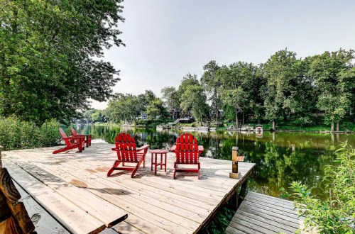 A serene lakeside dock with red chairs and a small table, surrounded by lush greenery and calm water.