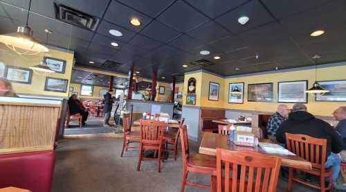 A cozy restaurant interior with wooden tables, chairs, and patrons dining. Walls adorned with framed pictures.