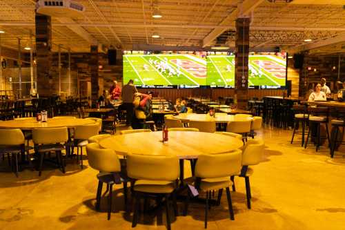 A spacious restaurant interior with large tables, chairs, and a big screen showing a football game.