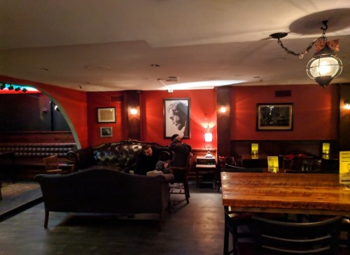 Cozy bar interior with dark wood furniture, warm lighting, and a large portrait on the wall. A person relaxes on a couch.