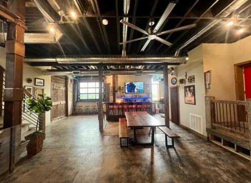 Interior of a spacious bar with a long wooden table, colorful lighting, and a large TV on the wall.