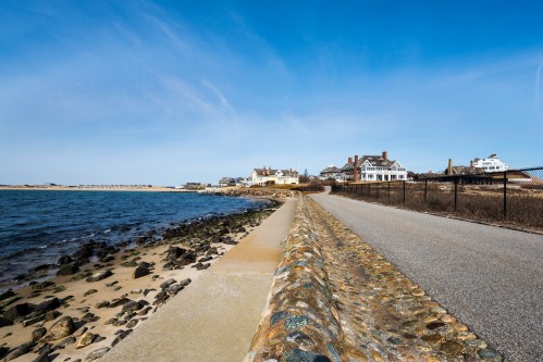 A coastal path lined with rocks, leading to beach houses under a clear blue sky.