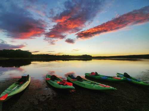 Colorful sunset over calm water with several green kayaks lined up on the shore.