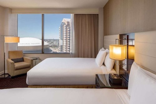 A modern hotel room with two beds, a chair, and a large window showcasing a city view.