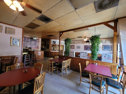Cozy restaurant interior with wooden walls, red tables, and hanging plants, featuring framed artwork and a ceiling fan.