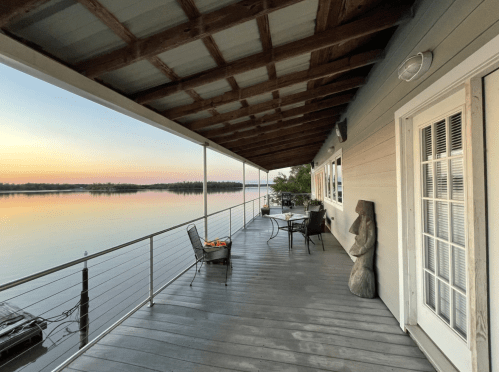 A serene lakeside porch with chairs, a table, and a wooden sculpture, overlooking calm waters at sunset.
