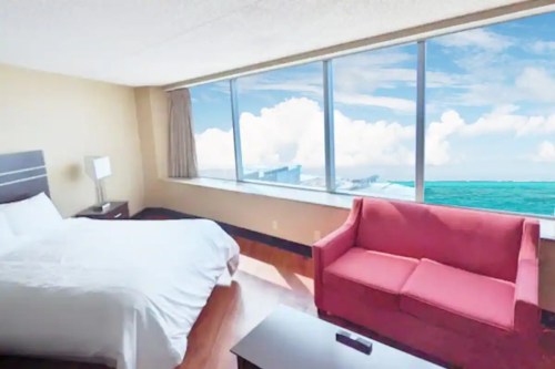 A bright hotel room with a bed and a pink sofa, featuring large windows overlooking a blue ocean and cloudy sky.