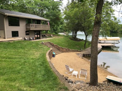 A lakeside property with a sandy beach, steps leading to the water, and a house surrounded by trees.