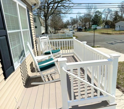 A porch with two rocking chairs, white railings, and a view of a quiet street lined with trees and houses.
