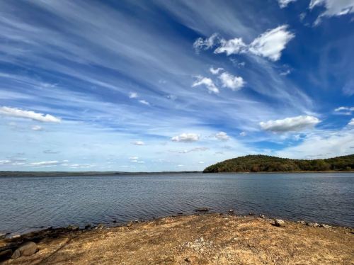 A serene lake scene with a sandy shore, gentle waves, and a backdrop of blue sky and wispy clouds.