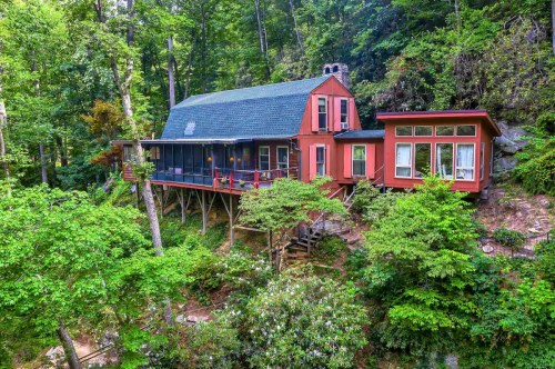 A charming red cabin nestled among lush green trees, featuring a porch and large windows, surrounded by nature.